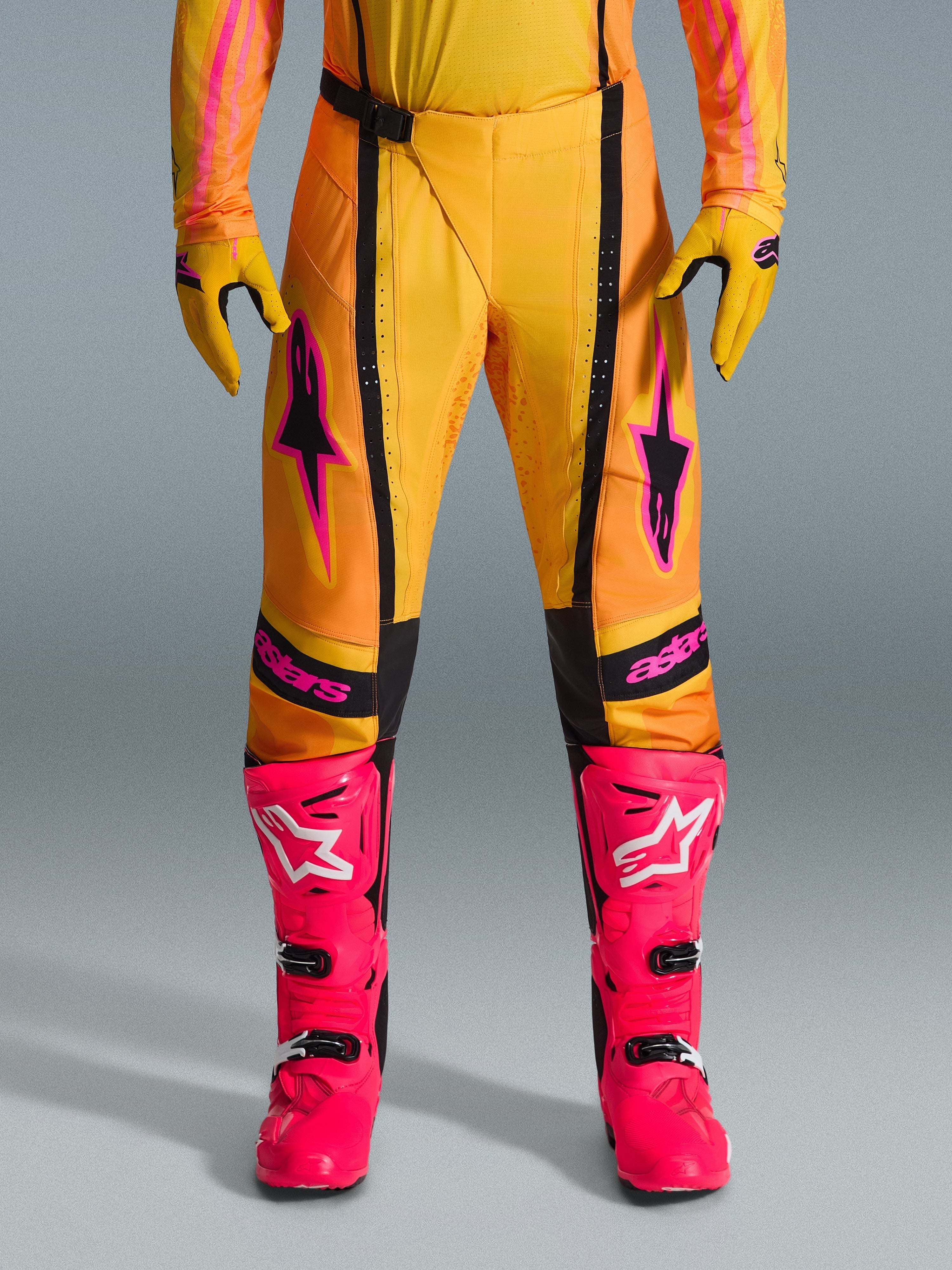 Alpinestars 2026 Techstar Nomur Hose, Motocross-Hose, Orange, Gelb und Pink, getragen mit passenden gelben Handschuhen und leuchtend pinkfarbenen Rennstiefeln, mit schwarzen Einsätzen und auffälligem pinkfarbenem Alpinestars-Branding, technische Offroad-Rennausrüstung
