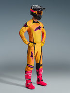 Motocross-Fahrer in Alpinestars 2026 Techstar Nomur Hose in Orange, Gelb und Pink, komplettes Rennbekleidungsset mit passendem Jersey und Handschuhen, leuchtend pinke Stiefel, schwarzer Helm mit orangefarbener Brille, Ganzkörperansicht vor grauem Hintergrund, professionelles Offroad-Rennoutfit