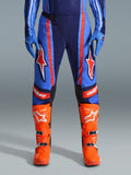 Alpinestars 2026 Techstar Nomur Hose, Motocross-Hose, Dunkelblau, Orange und UCLA-Blau, mit markanten orangefarbenen Streifen und großen weißen Stern-Logos auf den Oberschenkeln, getragen mit leuchtend orangefarbenen Rennstiefeln und blauen Handschuhen, athletische Rennpassform für Offroad-Performance