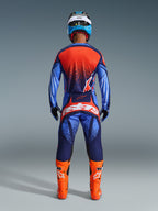 Motocross-Fahrer in der Rückansicht mit Alpinestars 2026 Techstar Nomur Hose in Dunkelblau, Orange und UCLA-Blau, kombiniert mit passendem Trikot und leuchtend orangefarbenen Stiefeln, professionelles Offroad-Rennausrüstungsset.