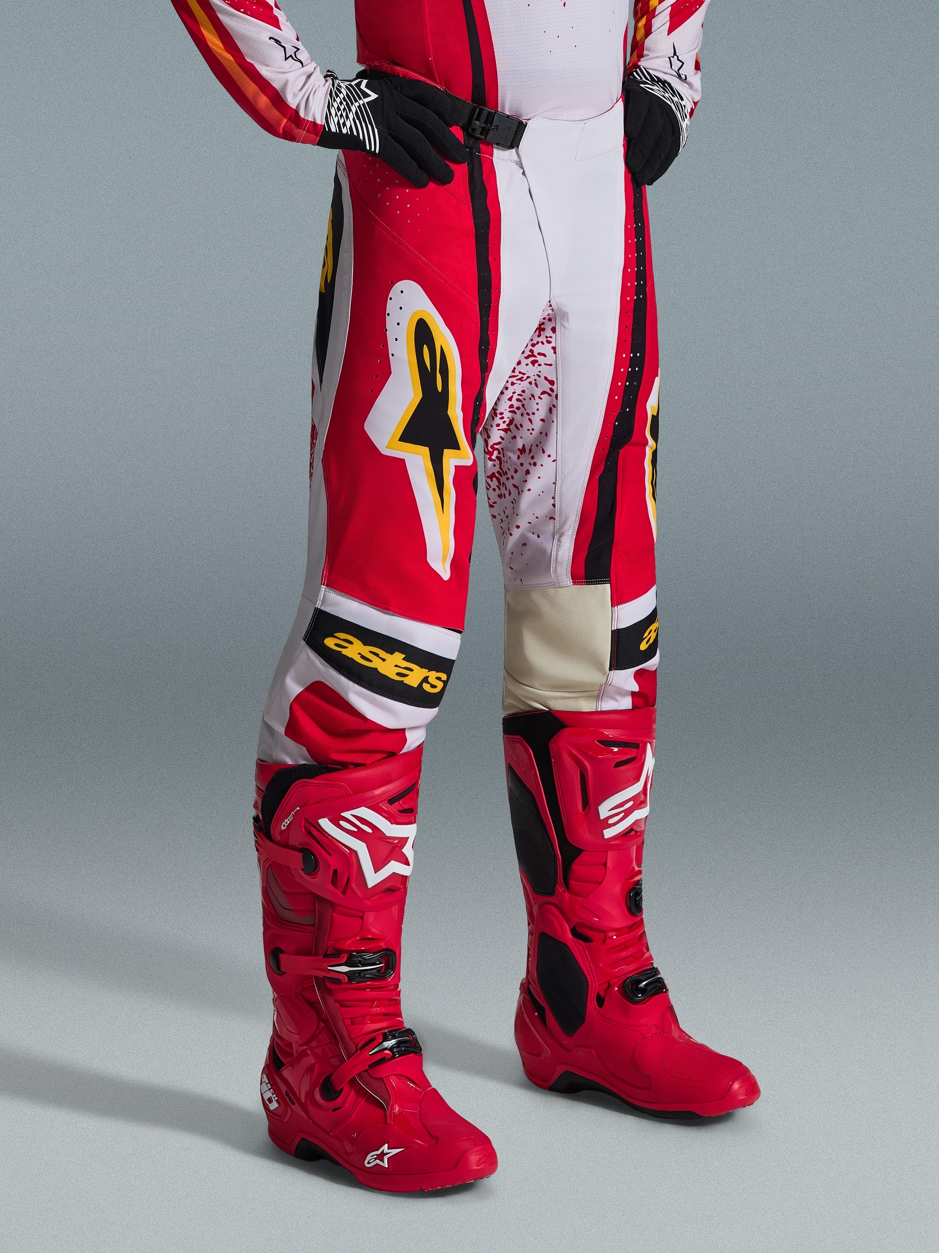 Alpinestars 2026 Techstar Nomur Hose, Motocross-Hose, Rot, Weiß und Schwarz mit gelben Akzenten, getragen mit passenden roten Motocross-Stiefeln und schwarzen Handschuhen, mit gelbem Alpinestars-Logo und „astars“-Branding auf den Knien, professionelle Offroad-Rennausrüstung.