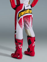 Rückansicht eines Motocross-Fahrers in der Alpinestars 2026 Techstar Nomur Hose in Rot und Weiß mit einem markanten gelben Logo am Gesäß, kombiniert mit leuchtend roten Tech 10 Motocross-Stiefeln, Studioaufnahme vor grauem Hintergrund.