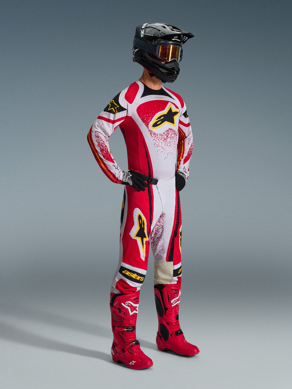 Alpinestars 2026 Techstar Nomur Hose, Motocross-Hose, Rot und Weiß mit schwarzen und gelben Akzenten, getragen von einem Fahrer mit passendem Renntrikot, leuchtend roten Stiefeln und einem schwarzen Helm mit goldener Brille, komplette professionelle Offroad-Wettbewerbsausrüstung