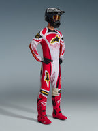 Alpinestars 2026 Techstar Nomur Hose, Motocross-Hose, Rot und Weiß mit schwarzen und gelben Akzenten, getragen von einem Fahrer mit passendem Renntrikot, leuchtend roten Stiefeln und einem schwarzen Helm mit goldener Brille, komplette professionelle Offroad-Wettbewerbsausrüstung