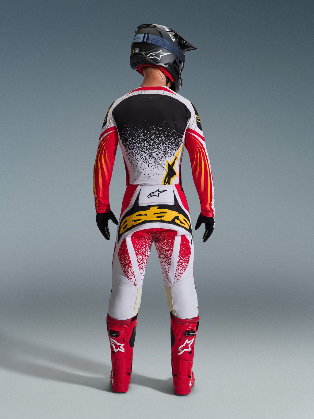Rückansicht eines Motocross-Fahrers im Alpinestars 2026 Techstar Nomur Trikot in Schwarz, Rot und Weiß mit gelben Akzenten, kombiniert mit einer passenden Motocross-Hose mit auffälligem gelbem Branding, leuchtend roten Rennstiefeln und einem schwarzen Helm; komplettes, aufeinander abgestimmtes Ausrüstungsset für den Offroad-Rennsport.
