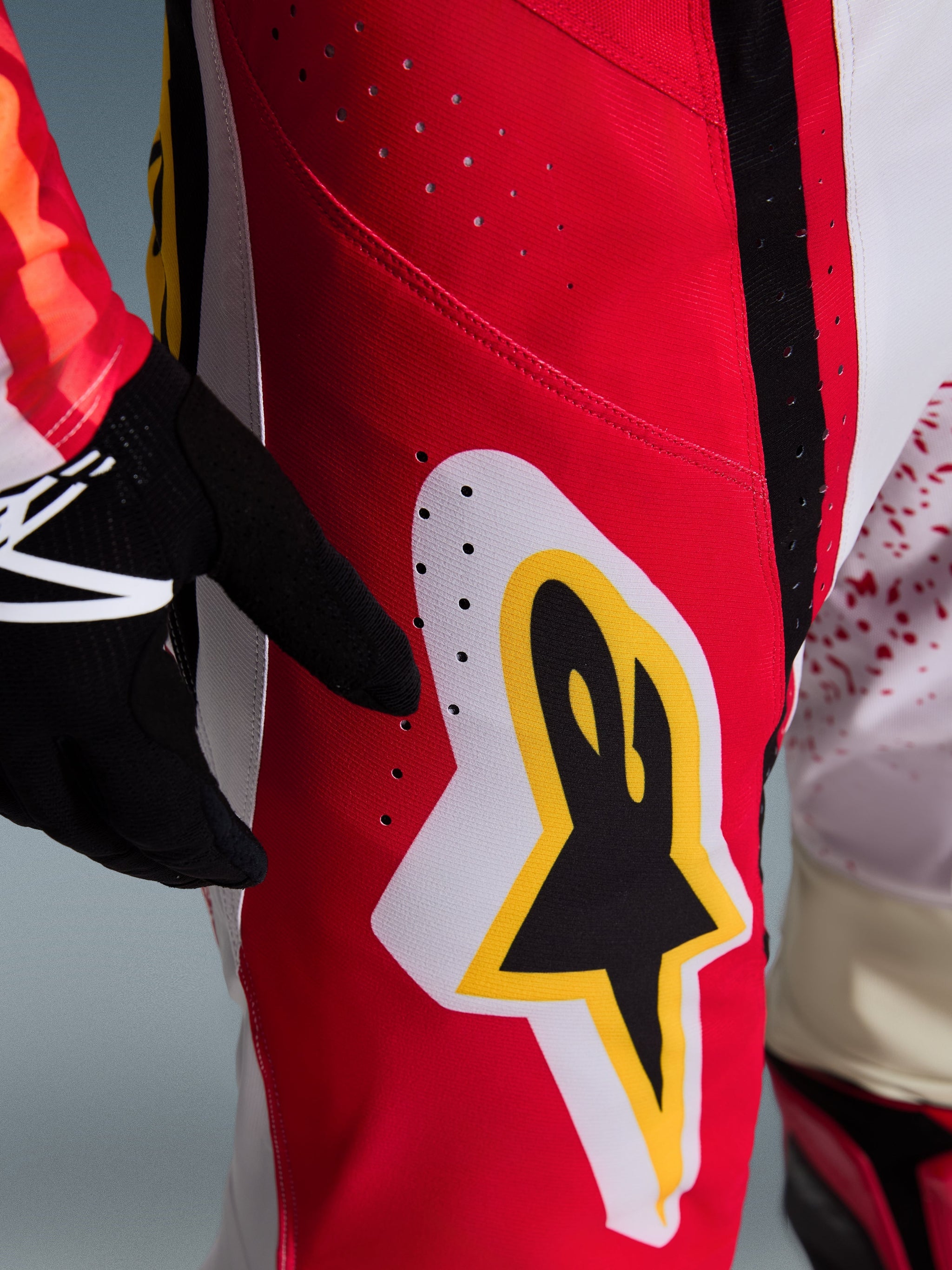 Alpinestars 2026 Techstar Nomur Hose, Motocross-Hose, Rot mit weißen und gelben Akzenten, Nahaufnahme des Oberschenkelbereichs mit lasergeschnittenen Belüftungslöchern und einem großen Stern-Logo, kombiniert mit schwarzen Handschuhen und einem bunten Trikot für Offroad-Performance.