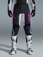 Alpinestars 2026 Fluid Grid Hose, Motocross-Hose, Schwarz mit lila und blauen Stern-Designs, kombiniert mit weißen Motocross-Stiefeln und schwarzen Handschuhen, mit verstärkten Einsätzen und einer sportlichen Offroad-Passform, grauer Hintergrund