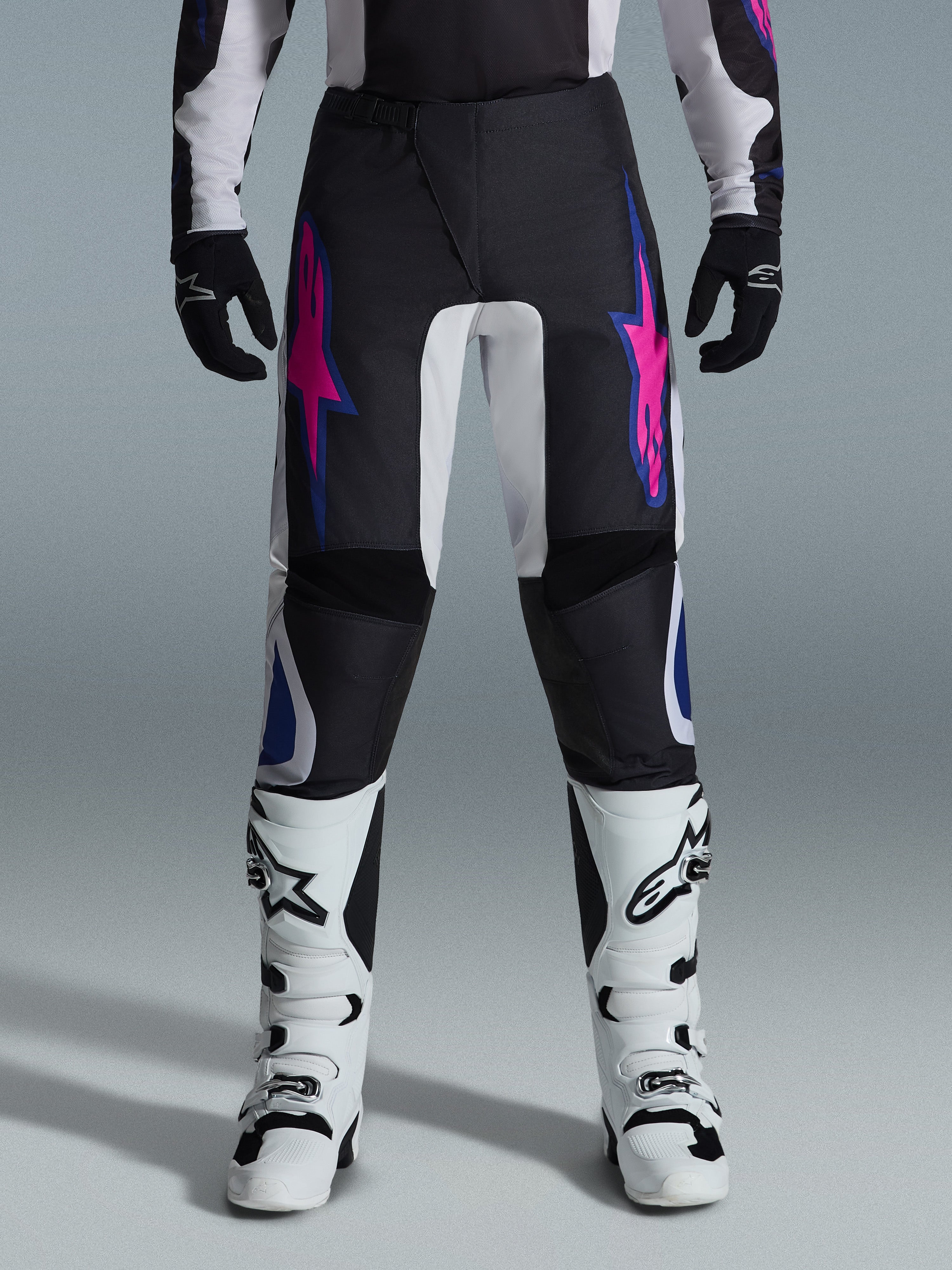 Alpinestars 2026 Fluid Grid Hose, Motocross-Hose, Schwarz mit lila und blauen Stern-Designs, kombiniert mit weißen Motocross-Stiefeln und schwarzen Handschuhen, mit verstärkten Einsätzen und einer sportlichen Offroad-Passform, grauer Hintergrund
