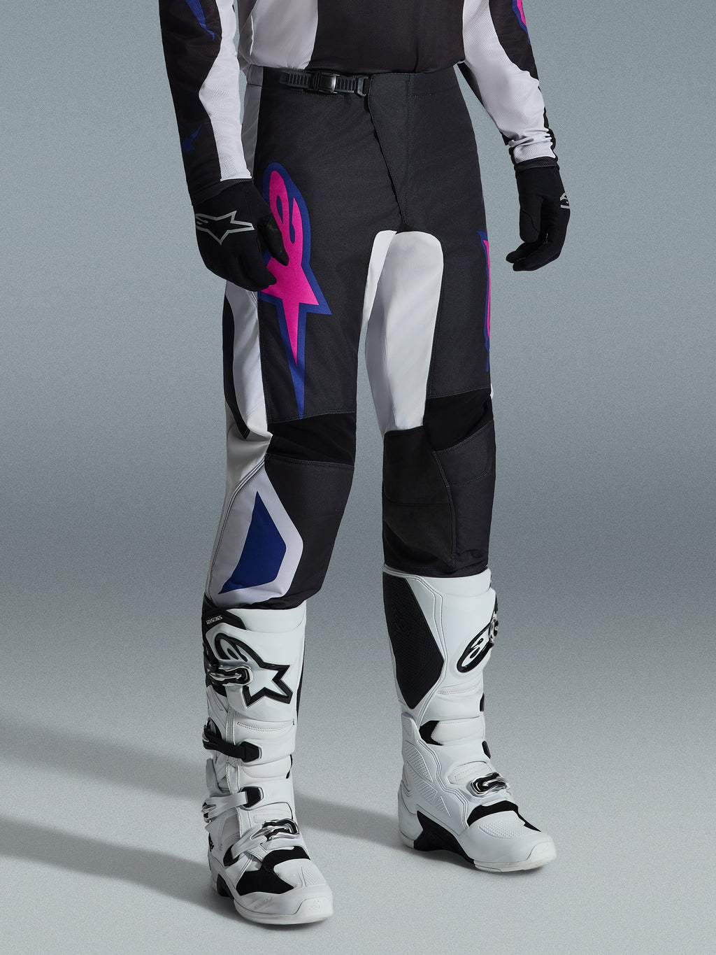 Alpinestars 2026 Fluid Grid Hose, Motocross-Hose, Schwarz, Hellgrau und Lila, mit einem auffälligen pinkfarbenen Stern-Logo und blauen geometrischen Akzenten, getragen mit weißen Motocross-Stiefeln und schwarzen Handschuhen, ergonomische Offroad-Fahrausrüstung