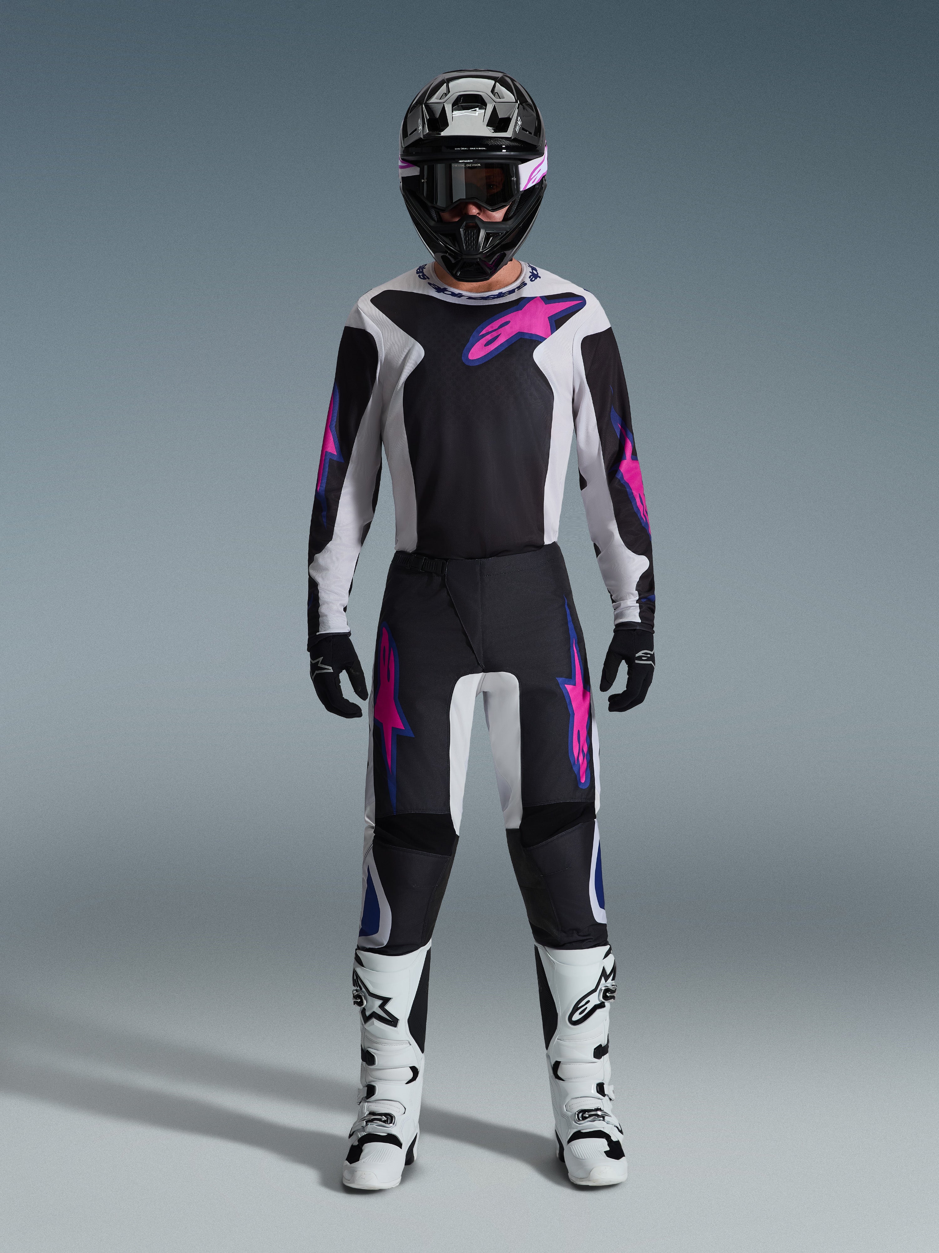 Motocross-Fahrer im Alpinestars 2026 Fluid Grid Ausrüstungsset, schwarz-hellgraues Jersey mit leuchtend pinkfarbenen Logos, schwarz-weiße Hose mit passenden pink-blauen Grafiken, schwarzer Helm mit pinkfarbener Brille, weiß-schwarze Stiefel, komplette Offroad-Rennausrüstung