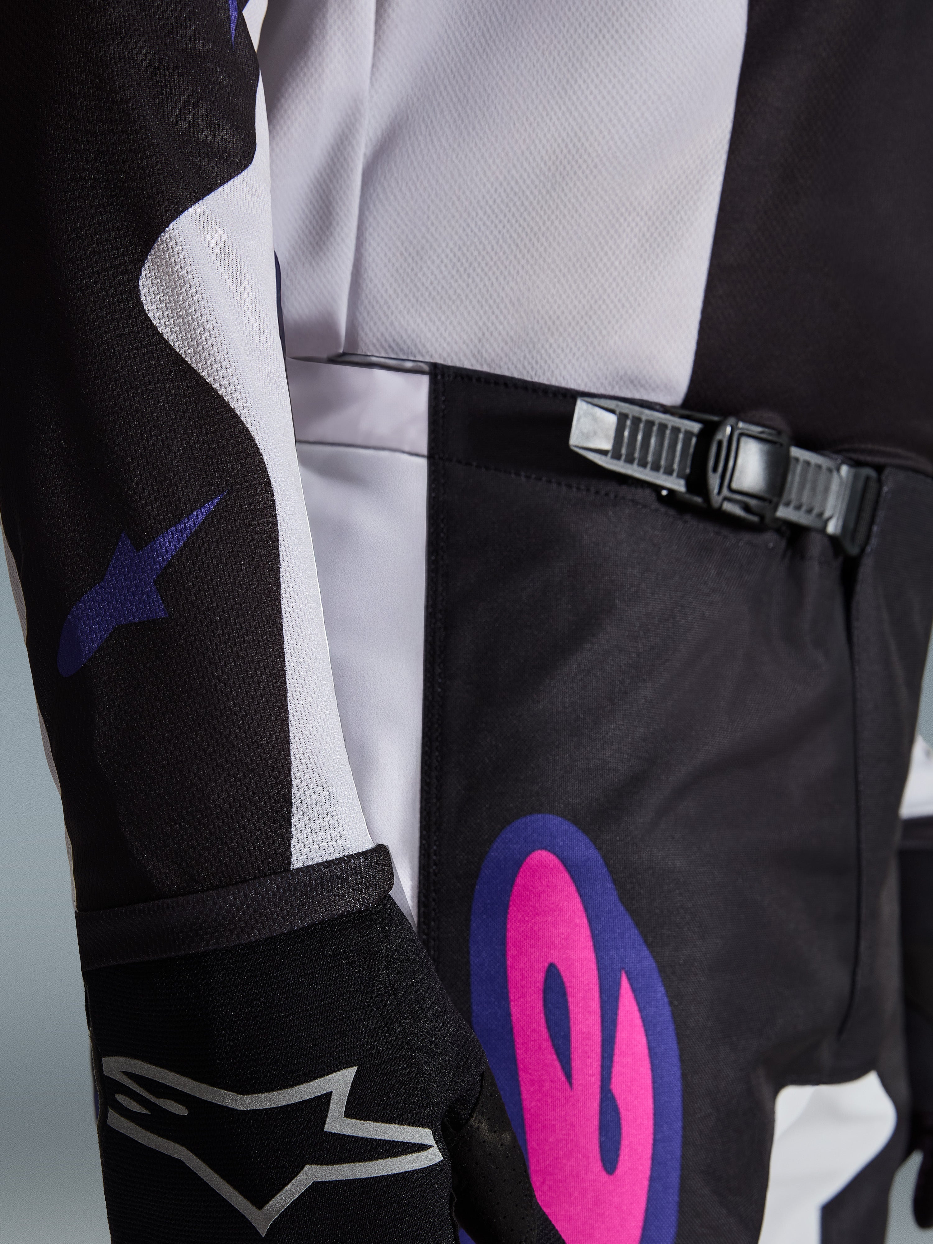 Alpinestars 2026 Fluid Grid Hose, Motocross-Hose, Schwarz, Hellgrau und Lila, Nahaufnahme mit dem Ratschenverschluss-System am Bund und einer auffälligen pink- und lilafarbenen Grafik an der Hüfte, getragen mit einem passenden Jersey und schwarzem Handschuh.