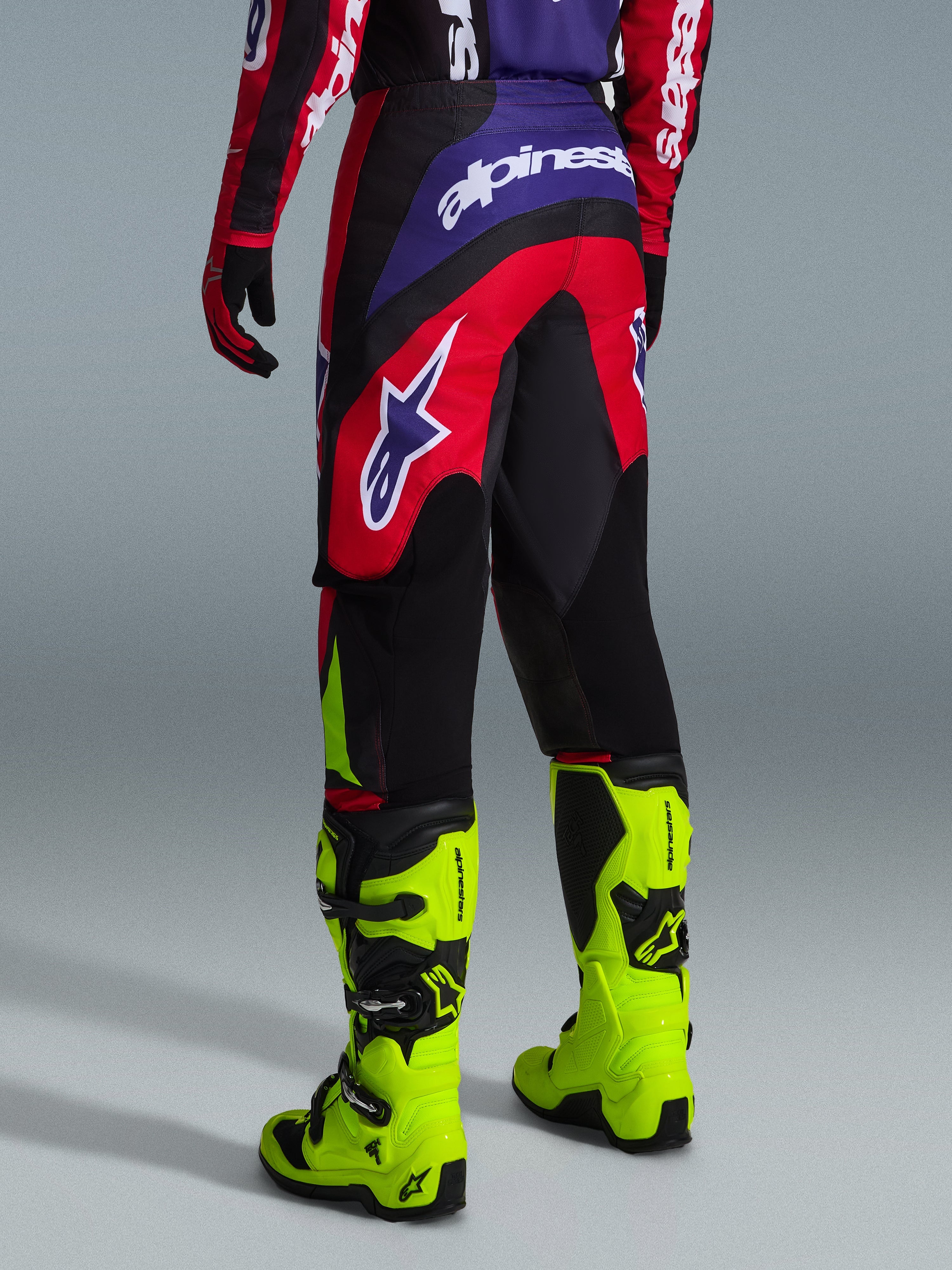 Alpinestars 2026 Fluid Grid Hose, Motocross-Hose, Rot, Schwarz und Lila, Rückansicht mit großem weißen Logo, getragen mit neongelben Rennstiefeln und passendem Jersey, strapazierfähiges Sport-Fit-Design für Offroad-Performance