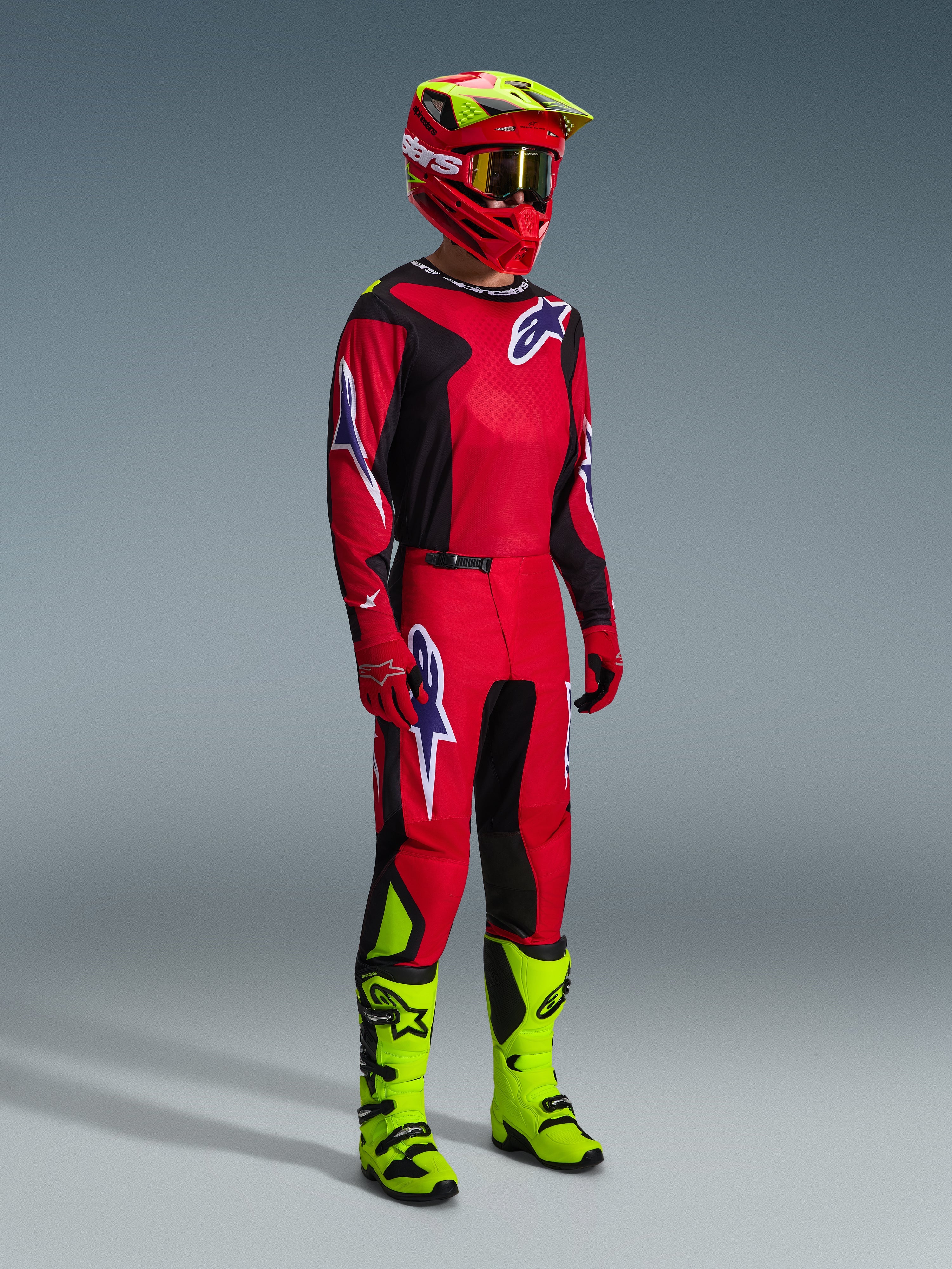 Motocross-Fahrer in der 2026 Fluid Grid Hose in Rot Schwarz Lila, komplette Rennausrüstung mit passendem Jersey, leuchtend roter und neongelber Helm und neongelbe Stiefel, abgestimmtes Offroad-Rennoutfit mit markantem Alpinestars-Branding