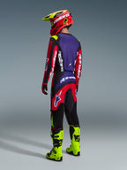 Alpinestars 2026 Fluid Grid Hose, Motocross-Hose, Rot, Schwarz und Lila, von einem Fahrer von hinten getragen mit passendem Trikot und neongelben Stiefeln, mit markantem Branding und ergonomischem Design für Offroad-Performance.