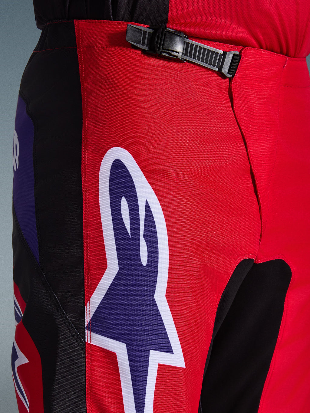 Alpinestars 2026 Fluid Grid Hose, Motocross-Hose, Rot, Schwarz und Lila, Nahaufnahme des Taillen- und Hüftbereichs mit einem schwarzen Ratschenverschluss und einem großen lila Alpinestars-Sternlogo mit weißer Umrandung, strapazierfähiges strukturiertes Gewebe für den Offroad-Rennsport.