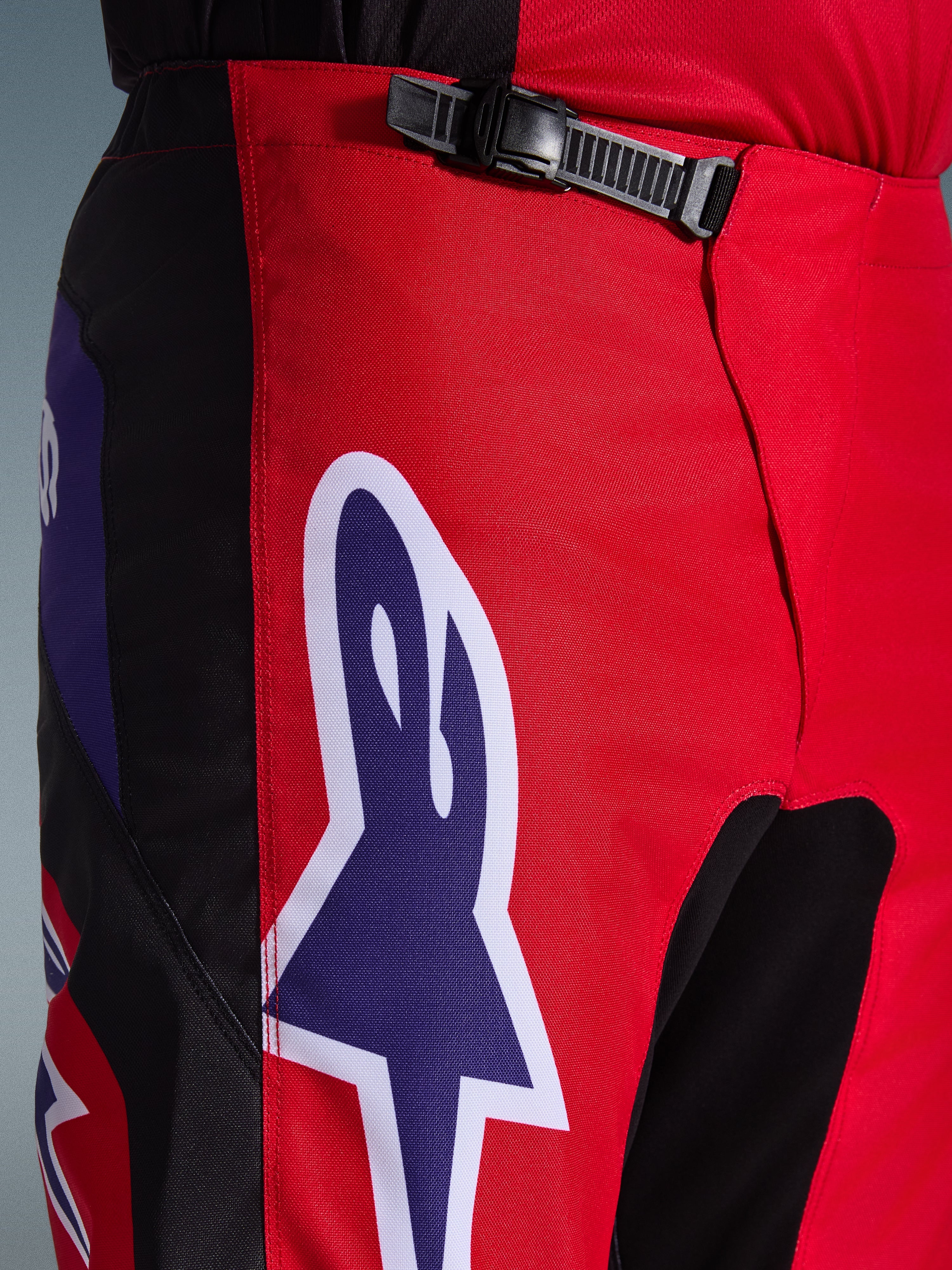 Alpinestars 2026 Fluid Grid Hose, Motocross-Hose, Rot, Schwarz und Lila, Nahaufnahme des Taillen- und Hüftbereichs mit einem schwarzen Ratschenverschluss und einem großen lila Alpinestars-Sternlogo mit weißer Umrandung, strapazierfähiges strukturiertes Gewebe für den Offroad-Rennsport.
