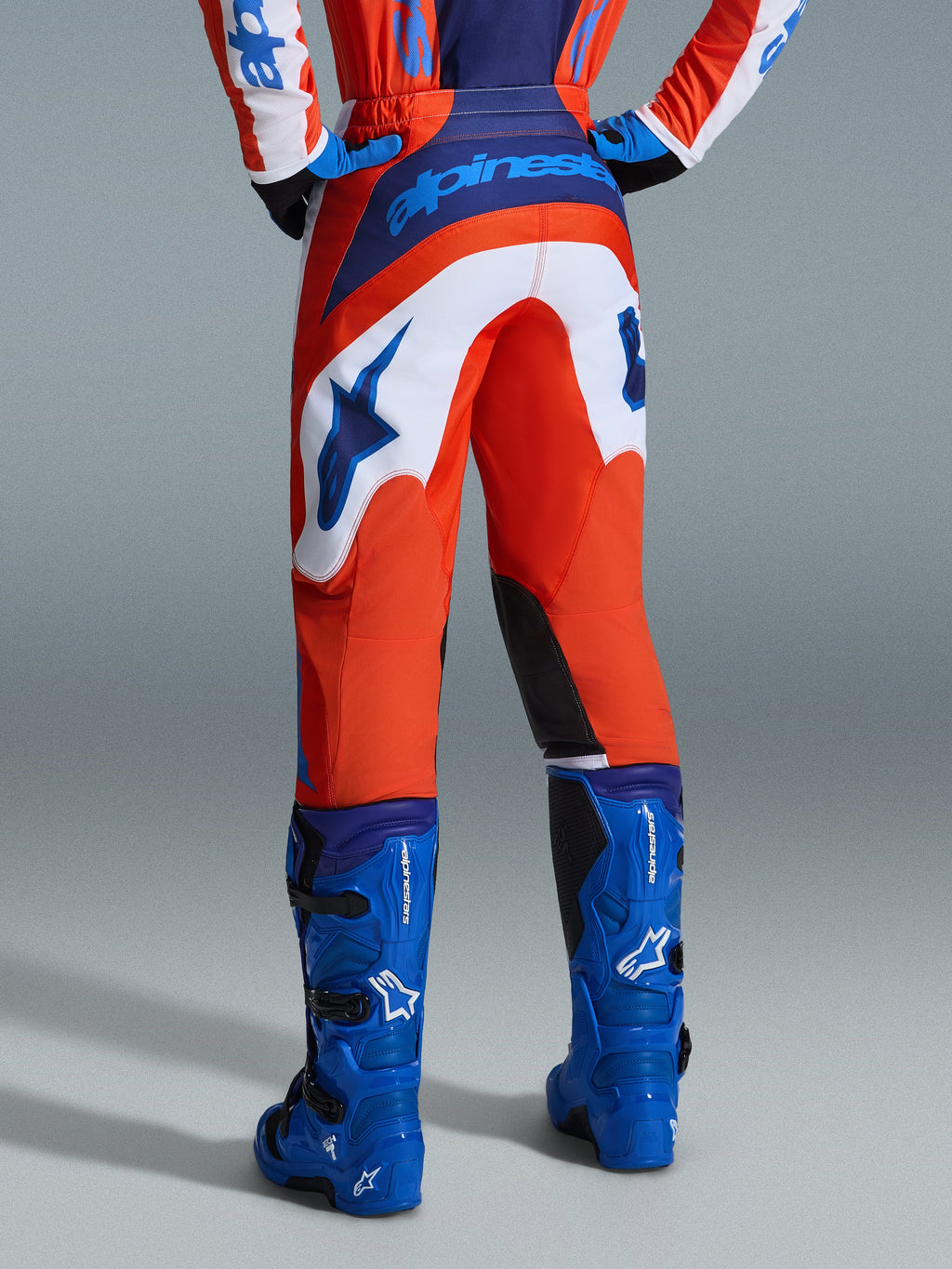 Alpinestars 2026 Fluid Grid Hose, Motocross-Hose, Orange und UCLA-Blau mit weißen Einsätzen, Rückansicht mit markantem blauem Branding auf der Gesäßpartie und Stern-Logo, kombiniert mit passenden blauen Motocross-Stiefeln, professionelle Offroad-Rennausrüstung