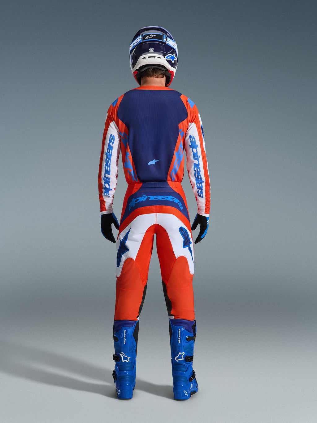 Alpinestars 2026 Fluid Grid Hose, Motocross-Hose, Orange und UCLA Blau, vollständige Rückansicht eines Fahrers in kompletter Rennausrüstung mit passendem Trikot, blauen Stiefeln und Helm, mit markantem Branding und ergonomischen Einsätzen für den Offroad-Sport.