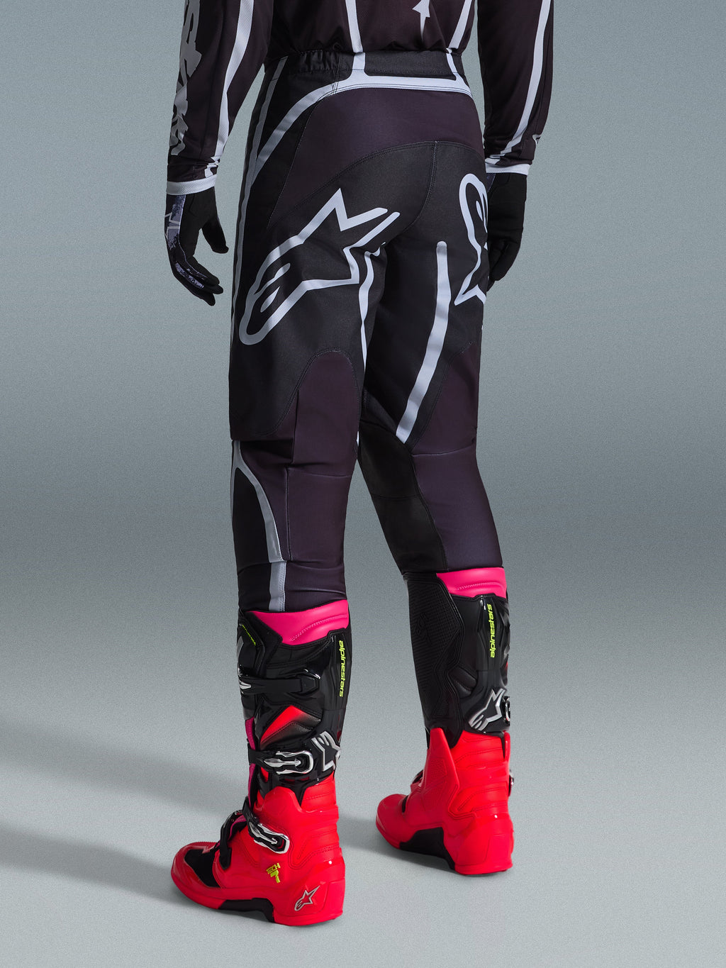 Alpinestars 2026 Fluid Apex Hose, Motocross-Hose, Schwarz und Grau, Rückansicht mit großen weißen Alpinestars-Logos am Gesäß, kombiniert mit leuchtend roten und pinken Stiefeln, strapazierfähige 600D-Polygewebe-Konstruktion für Offroad-Performance