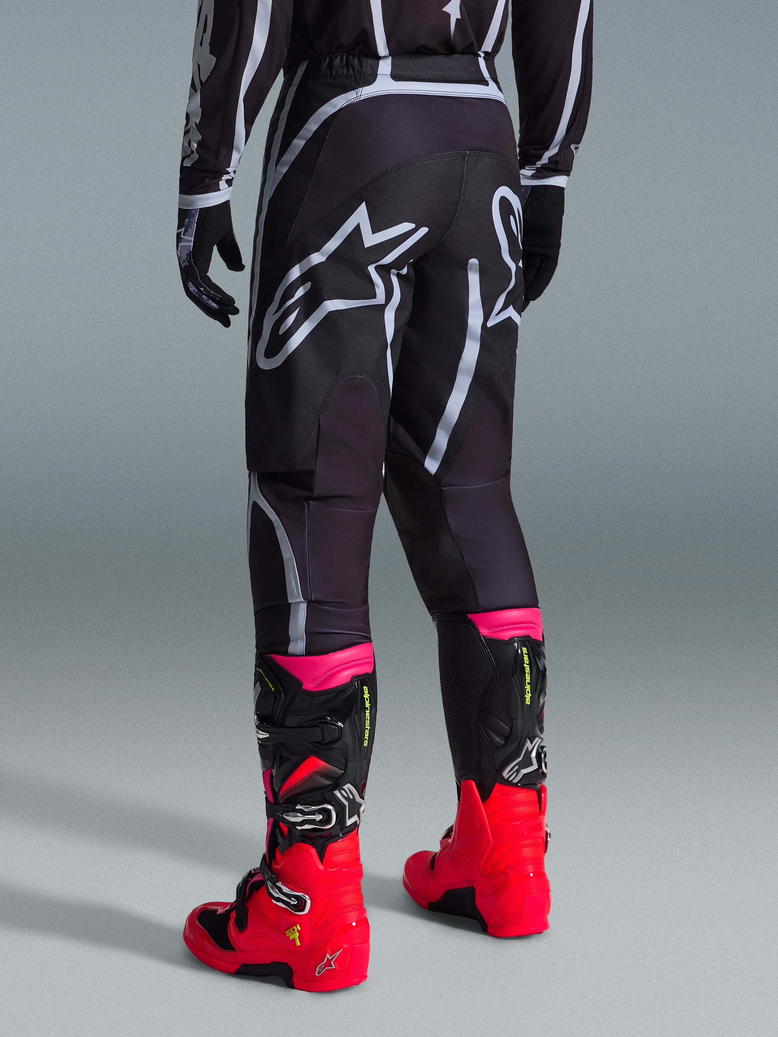 Alpinestars 2026 Fluid Apex Hose, Motocross-Hose, Schwarz und Grau, Rückansicht mit großen weißen Alpinestars-Logos am Gesäß, kombiniert mit leuchtend roten und pinken Stiefeln, strapazierfähige 600D-Polygewebe-Konstruktion für Offroad-Performance