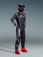 Alpinestars 2026 Fluid Apex Hose, Motocross-Hose, Schwarz und Grau, kombiniert mit passendem schwarz-weißem Trikot, schwarzem Helm mit pinker Brille und leuchtend roten Stiefeln, mit auffälligem weißem Branding an den Beinen und einem sportlichen Design für den Offroad-Rennsport.