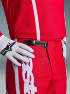 Alpinestars 2026 Fluid Apex Hose, Motocross-Hose, Rot und Weiß, Nahaufnahme des schwarzen Ratschenverschlusses am Bund, getragen mit weißen Alpinestars Handschuhen und passendem rotem Jersey, strapazierfähige Performance-Ausrüstung für den Offroad-Sport.