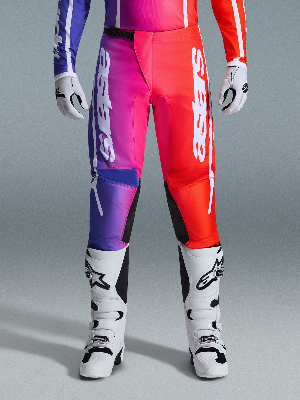 Alpinestars 2026 Fluid Apex Hose, Motocross-Hose, mehrfarbiges Design mit violettem, pinkem und rotem Farbverlauf, großes weißes Alpinestars-Branding auf den Beinen, kombiniert mit weiß-schwarzen Motocross-Stiefeln und Handschuhen, professionelle Offroad-Rennausrüstung.