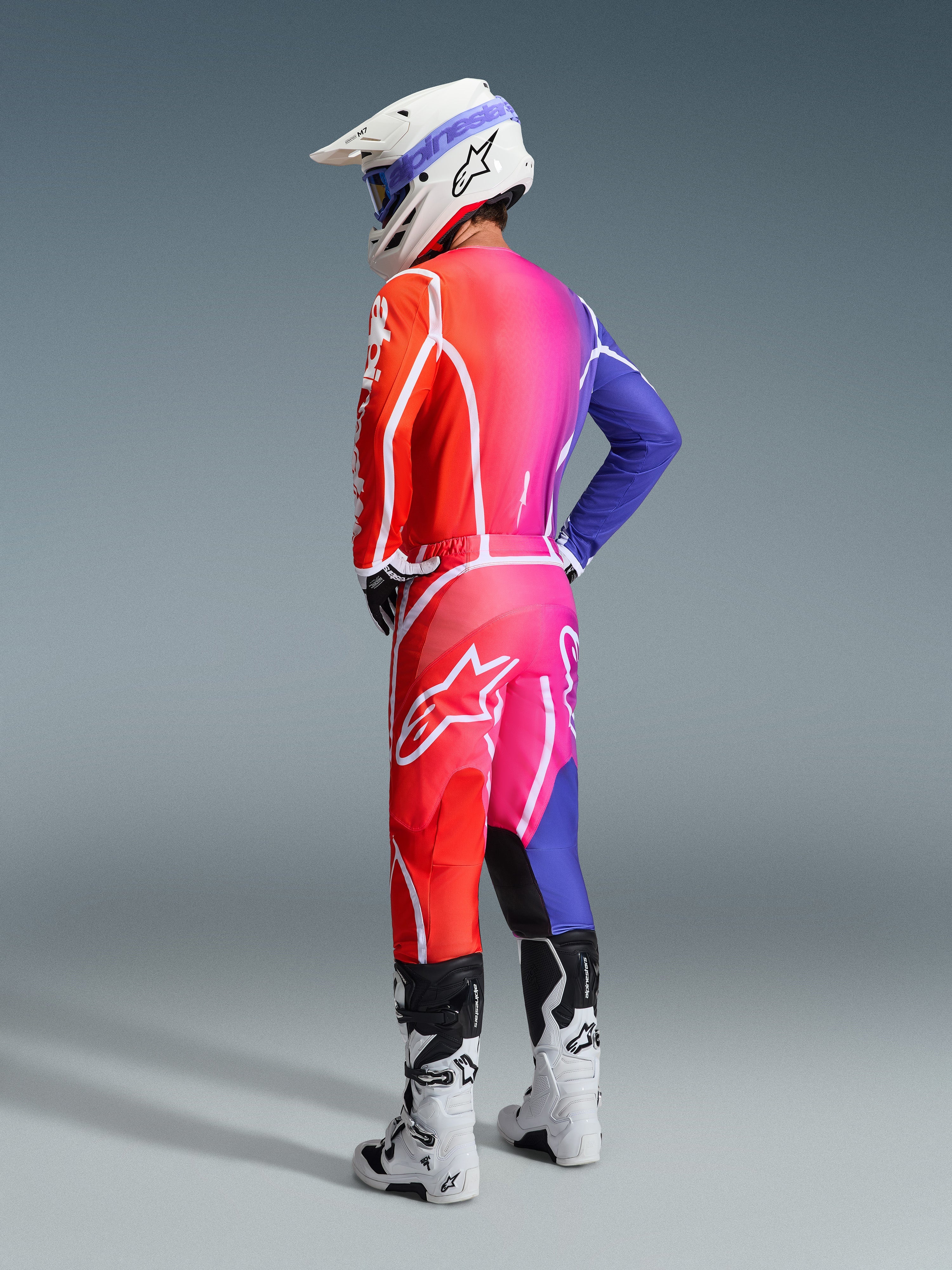 Motocross-Fahrer in Alpinestars 2026 Fluid Apex Ausrüstung, mehrfarbiges Trikot und Hose mit einem Farbverlauf in Rot, Pink und Violett, weiße Logos, weißer Helm und weiße Stiefel, koordiniertes Outfit für den Offroad-Rennsport
