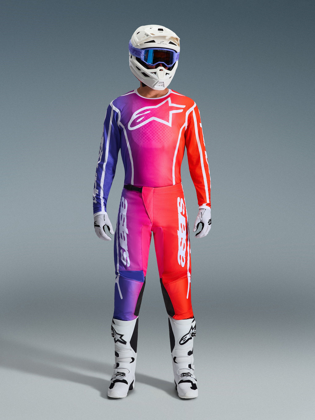 Motocross-Fahrer in 2026 Fluid Apex Hose und passendem Jersey, lila und orangefarbener Farbverlauf, weißer Helm mit Goggles, weiße Stiefel und Handschuhe, abgestimmte Alpinestars Rennausrüstung für Offroad-Performance.
