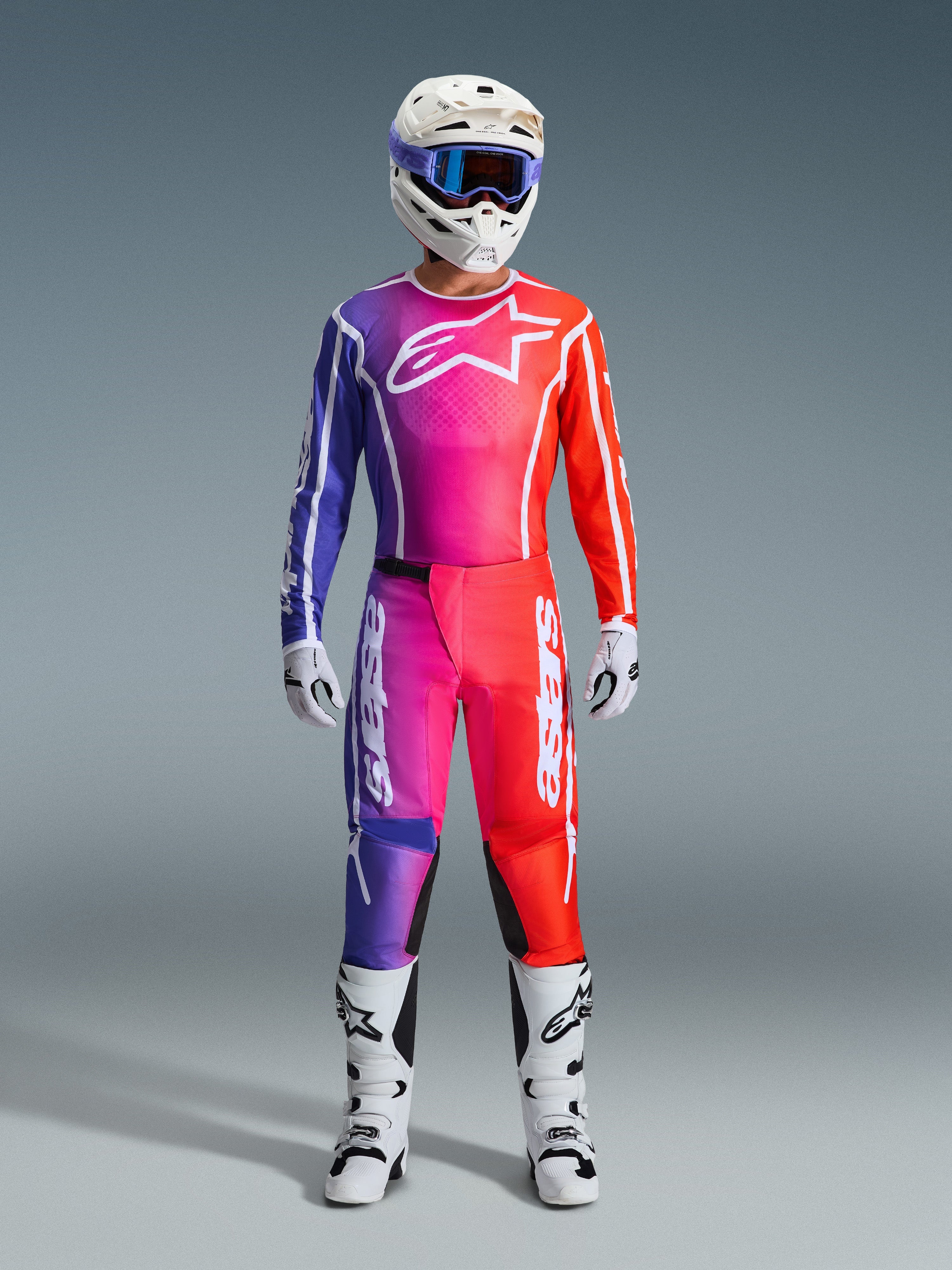Motocross-Fahrer in 2026 Fluid Apex Hose und passendem Jersey, lila und orangefarbener Farbverlauf, weißer Helm mit Goggles, weiße Stiefel und Handschuhe, abgestimmte Alpinestars Rennausrüstung für Offroad-Performance.