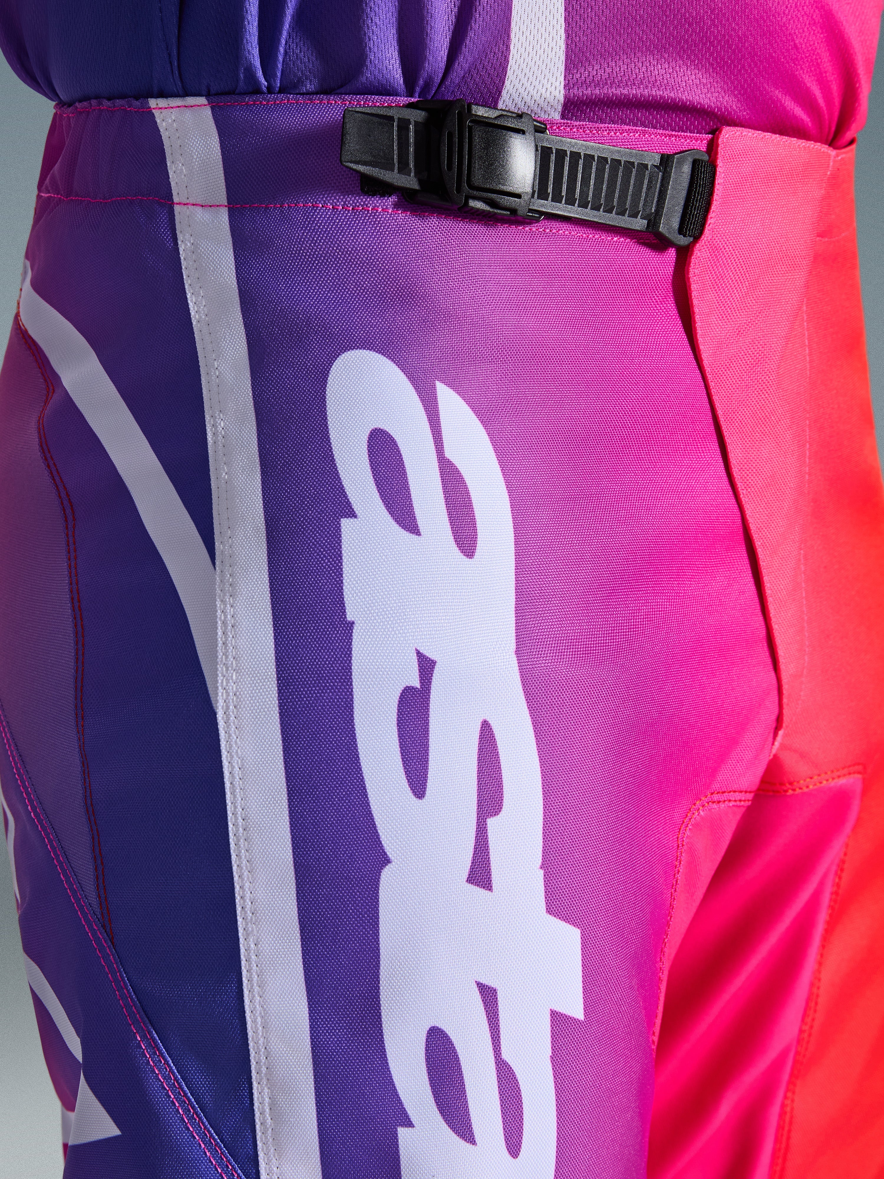 Alpinestars 2026 Fluid Apex Hose, Motocross-Hose, Mehrfarbig Lila, Pink und Rot, Nahaufnahme des Taillen- und Hüftbereichs mit schwarzem Ratschenverschluss-System und großem weißen vertikalen Alpinestars-Logo, lebendiges Farbverlauf-Design mit strapazierfähiger Polygewebe-Struktur