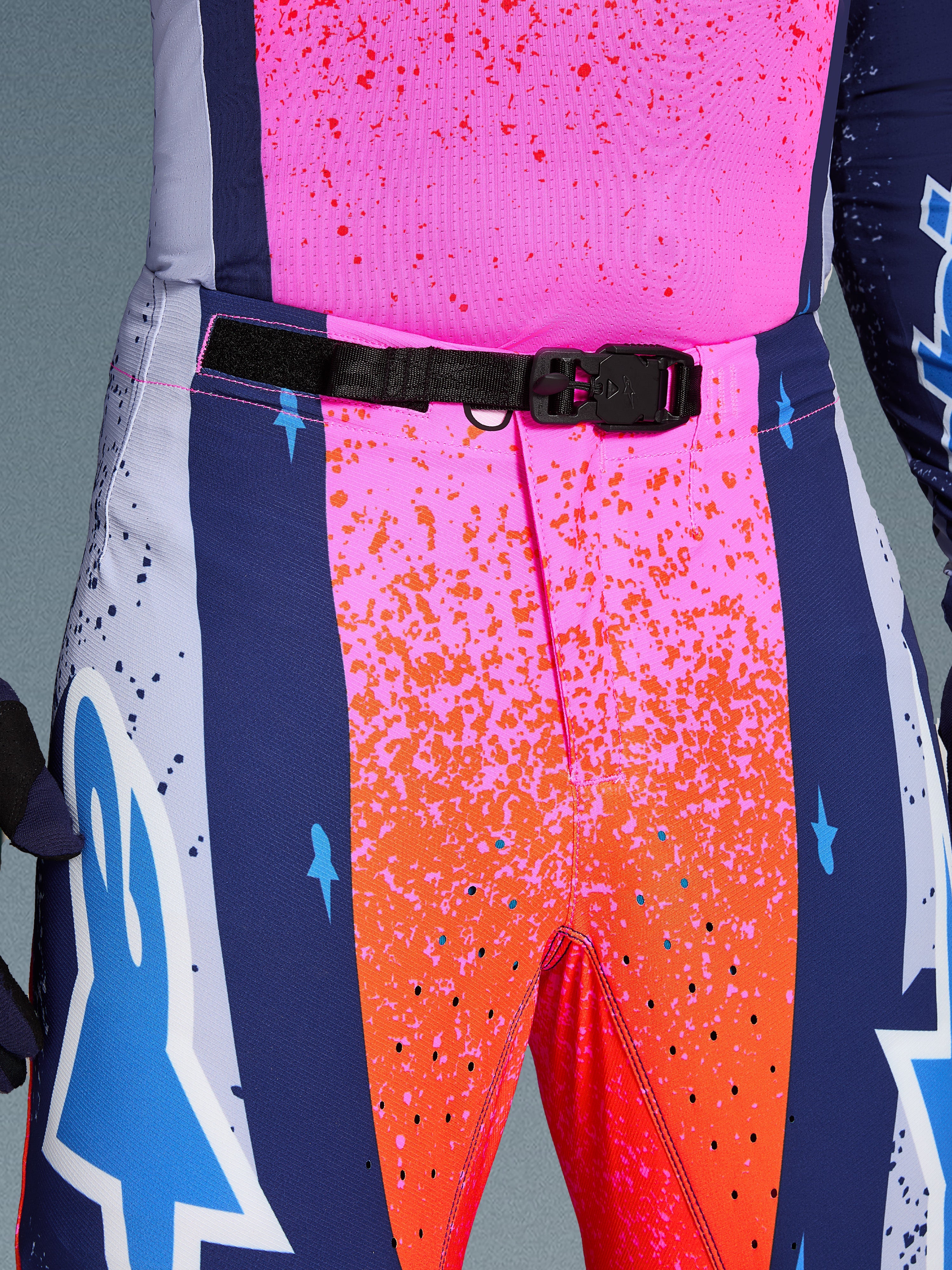 Alpinestars Limited Edition Supertech Records Hose, Motocross-Hose, Mehrfarbig Pink und Orange mit marineblauen Seiteneinsätzen, Nahaufnahme des Hosenbundes mit schwarzer, selbstverriegelnder SX1-Magnetschnalle, Ripstop-Gewebe mit Splatter-Grafiken und lasergeschnittenen Perforationen.