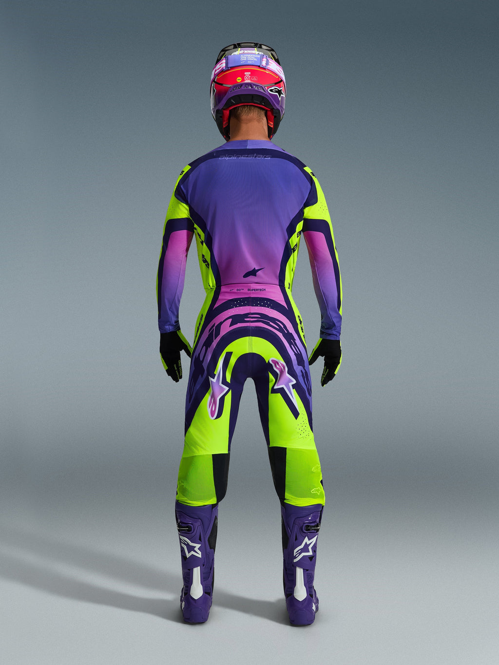 Motocross rider from behind wearing the 2026 Supertech Vista Jersey and matching pants, vibrant yellow and purple design with pink gradients, purple boots with white Alpinestars logos, black gloves, and a red and purple racing helmet, professional-grade racing gear set.