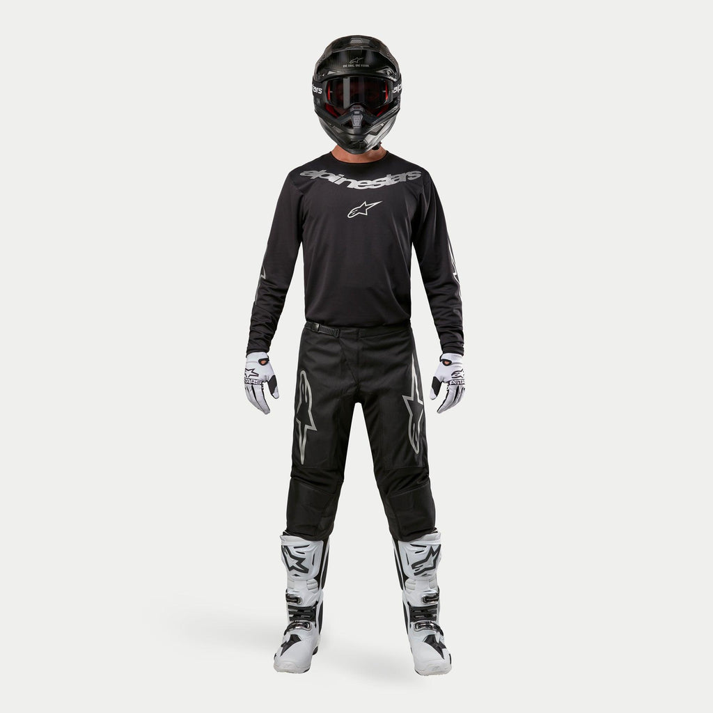 Motocross rider wearing complete racing gear, Alpinestars 2024 Fluid Graphite Pants in Black and Silver, black long-sleeve jersey with silver branding, black helmet with goggles, white and black boots, white and black gloves, coordinated outfit for motocross
