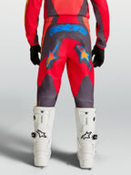 Alpinestars Jugend Fluid Hunt Hose, Motocrosshose in Rot und Grau, Rückansicht mit blauem astars-Branding und Sternengrafiken, strapazierfähiges 600D-Polygewebe, kombiniert mit weißen MX-Stiefeln.