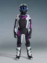 Youth motocross rider wearing Alpinestars 2026 Youth Fluid Grid Pants in Black, Light Gray, and Purple, matching long-sleeve jersey, black helmet with pink and purple goggles, white and black boots, and black gloves, full coordinated gear set for off-road riding.