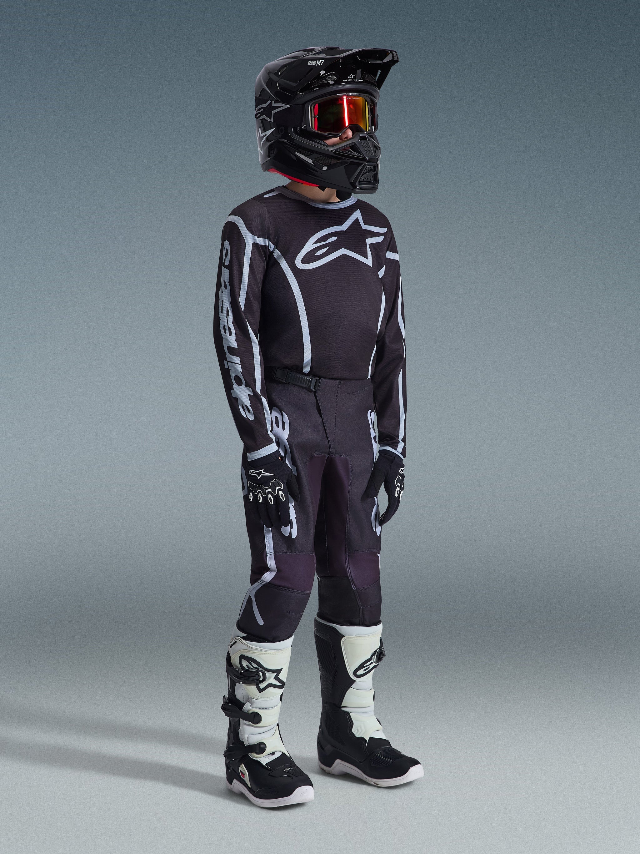 Alpinestars 2026 Jugend Fluid Apex Hose, Motocross-Hose, Schwarz und Grau, kombiniert mit passendem schwarz-weißem Trikot, schwarzem Helm und weiß-schwarzen Stiefeln, markantes Alpinestars-Branding und grafische Linien, abgestimmtes Rennoutfit für Jugend-Motocross