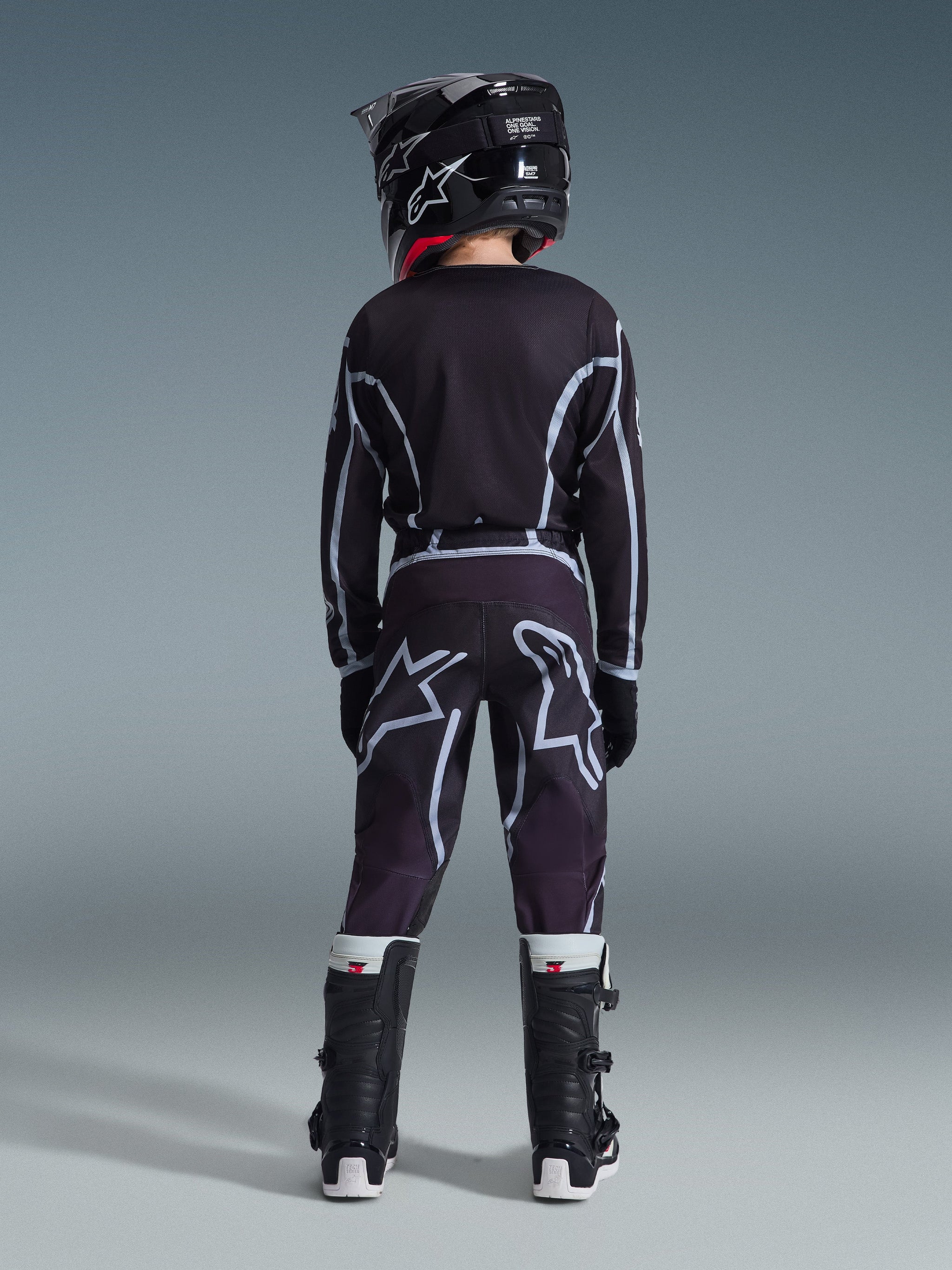 Rückansicht eines jugendlichen Motocross-Fahrers, der die Alpinestars 2026 Jugend Fluid Apex Hose in Schwarz und Grau, ein passendes Langarmtrikot, einen schwarzen Helm und schwarz-weiße Motocross-Stiefel trägt, abgestimmte Offroad-Rennausrüstung.