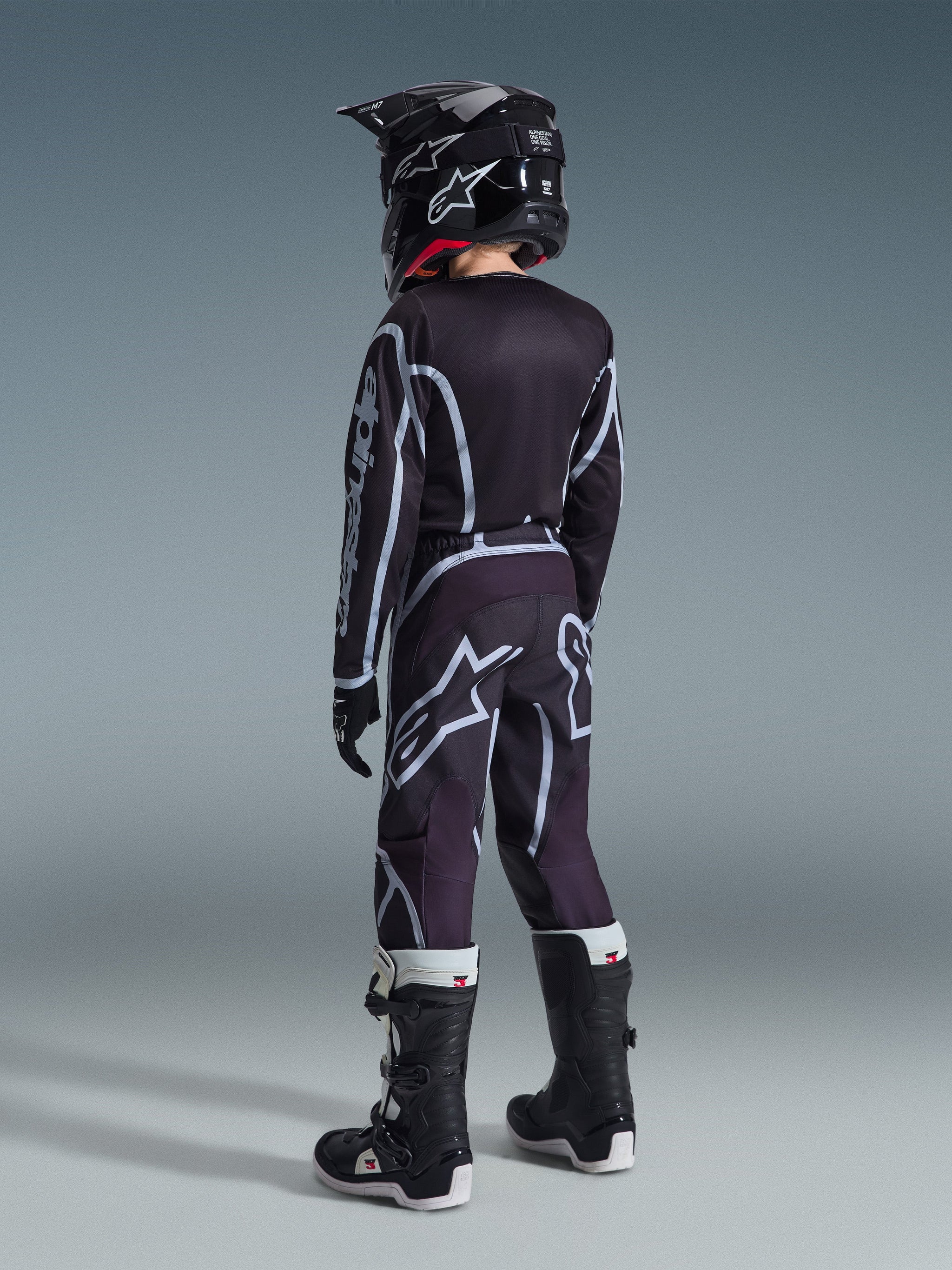 Alpinestars 2026 Youth Fluid Apex Hose, Jugend-Motocross-Hose, Schwarz und Grau, Rückansicht eines jungen Fahrers mit passendem schwarzem Trikot und Helm, auffällige weiße Logos und geometrische Muster, kombiniert mit schwarz-weißen Stiefeln für den Offroad-Rennsport.