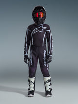 Jugend-Motocross-Fahrer in kompletter Rennausrüstung, 2026 Jugend Fluid Apex Hose in Schwarz und Grau, passendes schwarzes Trikot mit weißem Alpinestars-Logo, schwarzer Helm mit rot verspiegelter Brille, weiß-schwarze Stiefel, schwarz-weiße Handschuhe, abgestimmtes Outfit für den Offroad-Rennsport