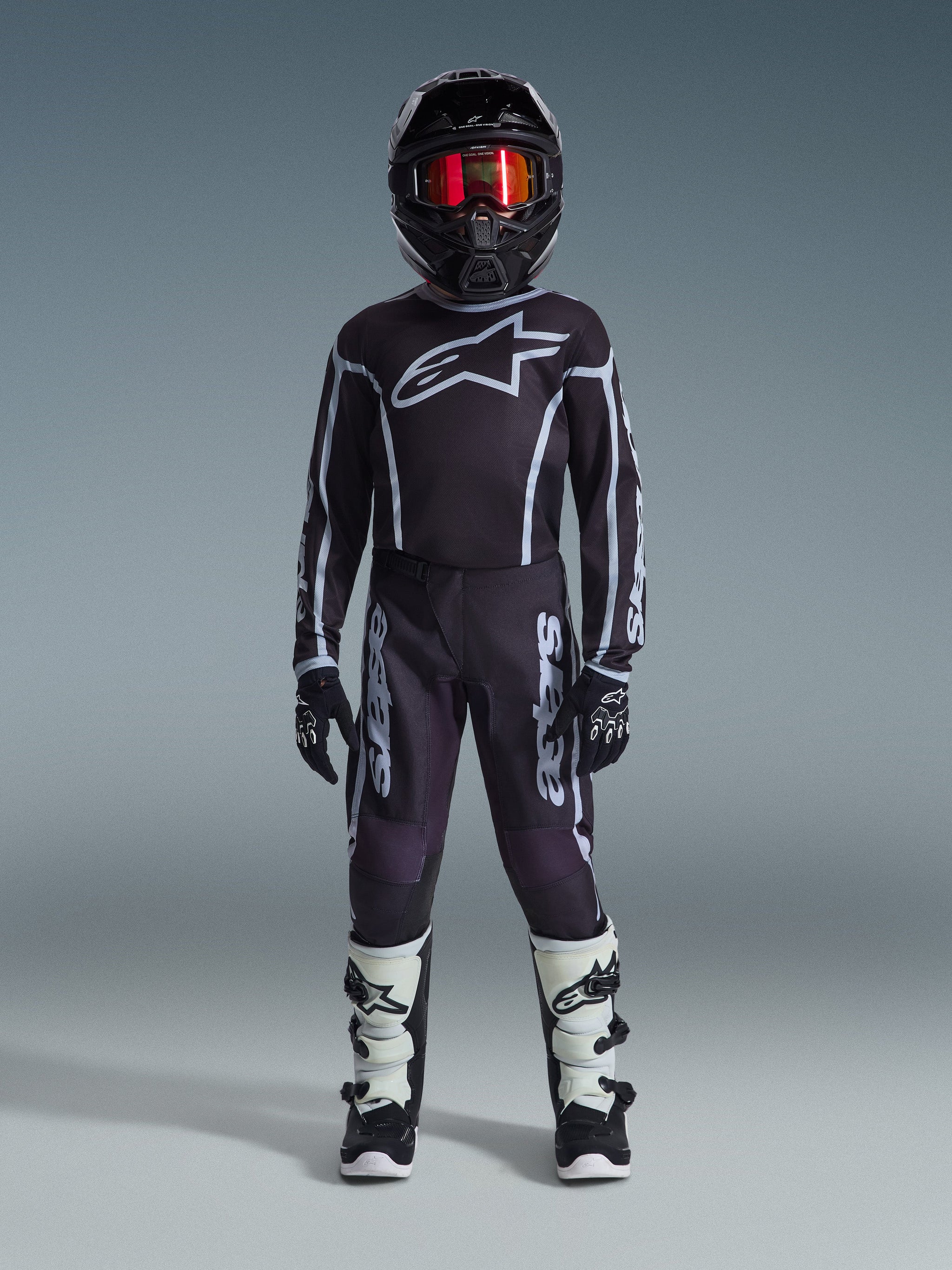 Jugend-Motocross-Fahrer in kompletter Rennausrüstung, 2026 Jugend Fluid Apex Hose in Schwarz und Grau, passendes schwarzes Trikot mit weißem Alpinestars-Logo, schwarzer Helm mit rot verspiegelter Brille, weiß-schwarze Stiefel, schwarz-weiße Handschuhe, abgestimmtes Outfit für den Offroad-Rennsport