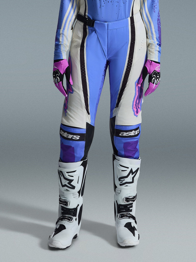 Alpinestars 2026 Stella Techstar Nomur Damen-Hose, Motocross-Hose, Hellblau, Off-White und Schwarz, getragen mit weißen Motocross-Stiefeln und pink-schwarzen Handschuhen, mit „astars“-Branding auf den Knien und technischem Mehrbahnen-Design für Fahrerinnen.