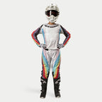 Motocross-Fahrerin in Alpinestars Women Stella 2024 Techstar Hose, Schwarz Multicolor mit weißer Basis sowie roten, gelben und blauen Streifen, passendes Langarmtrikot, weißer Helm und schwarze Stiefel, komplette Rennausrüstung für Offroad-Wettbewerbe