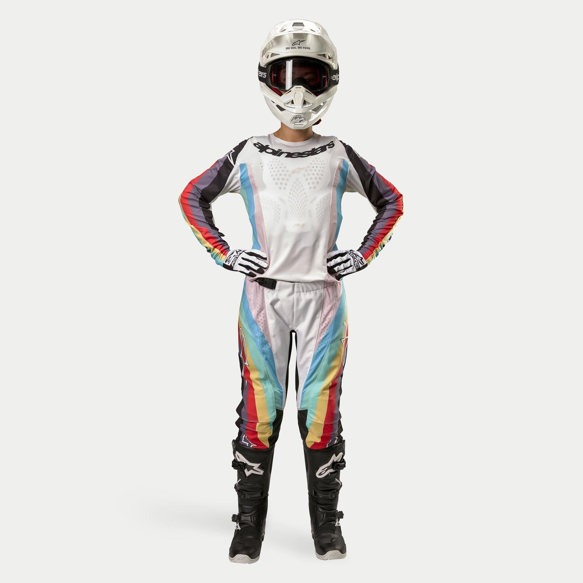 Motocross-Fahrerin in Alpinestars Women Stella 2024 Techstar Hose, Schwarz Multicolor mit weißer Basis sowie roten, gelben und blauen Streifen, passendes Langarmtrikot, weißer Helm und schwarze Stiefel, komplette Rennausrüstung für Offroad-Wettbewerbe
