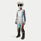 Motocross-Fahrerin mit Alpinestars Stella 2024 Techstar Hose, Motocross-Hose, Schwarz Mehrfarbig mit weißen Einsätzen und lebhaften diagonalen Streifen, passendem Trikot, weißem Helm und schwarzen Stiefeln, koordiniertes Offroad-Rennoutfit für Damen