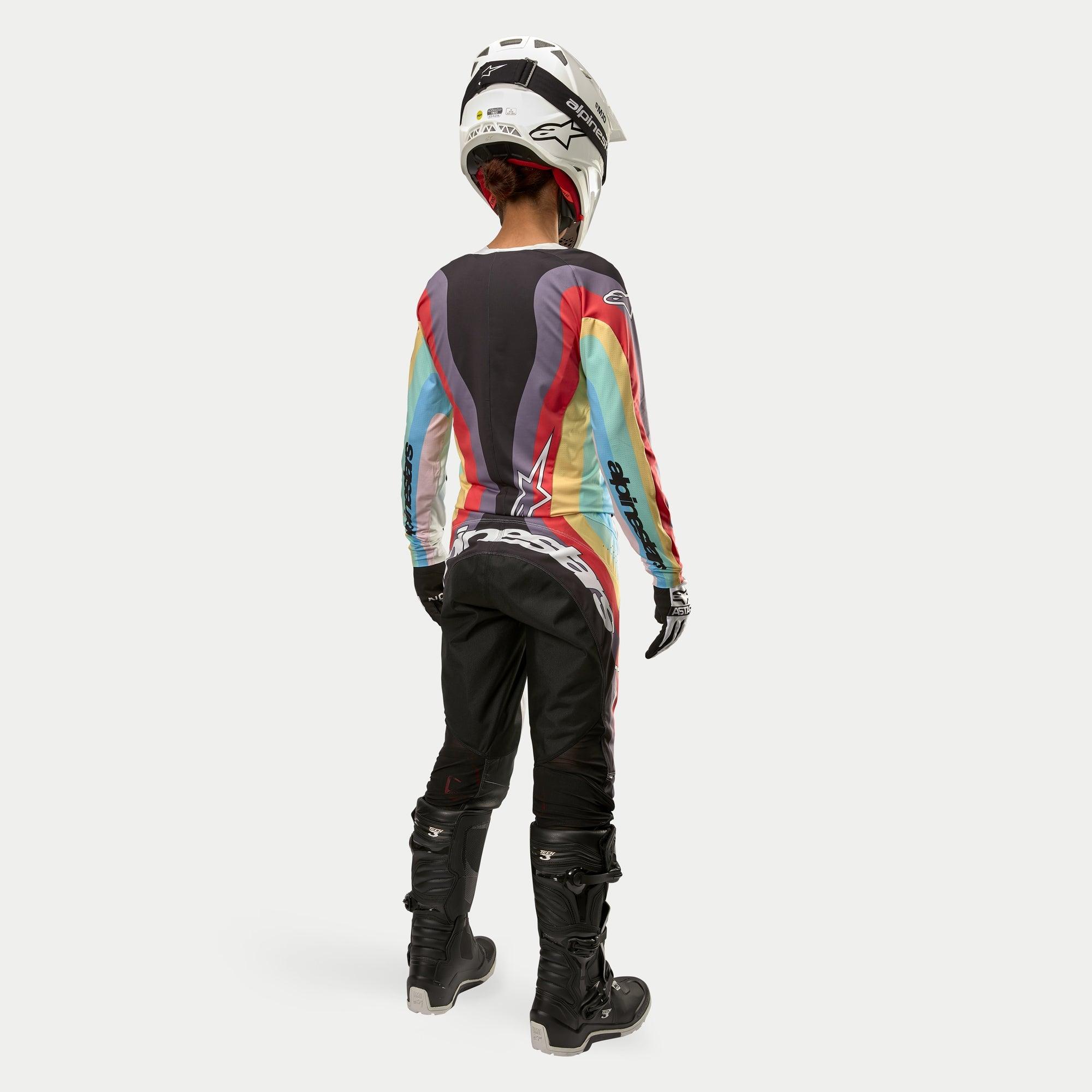 Motocross-Fahrerin von hinten in Alpinestars Stella Techstar Hose 2024 in Schwarz-Mehrfarbig, mit großem weißem Branding am Gesäß, kombiniert mit einem bunt gestreiften Trikot, weißem Helm und schwarzen Tech 3 Stiefeln für den Offroad-Rennsport.