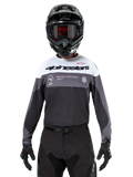Motocross rider wearing the Alpinestars Pro-Dura Jersey in Black Magnet White, long-sleeve performance top with white and grey color blocks, worn with a black helmet and goggles, featuring 'alpinestars' branding and 'ONE GOAL ONE VISION' slogan, off-road motorcycle racing apparel