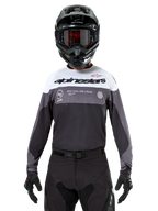 Motocross rider wearing the Alpinestars Pro-Dura Jersey in Black Magnet White, long-sleeve performance top with white and grey color blocks, worn with a black helmet and goggles, featuring 'alpinestars' branding and 'ONE GOAL ONE VISION' slogan, off-road motorcycle racing apparel