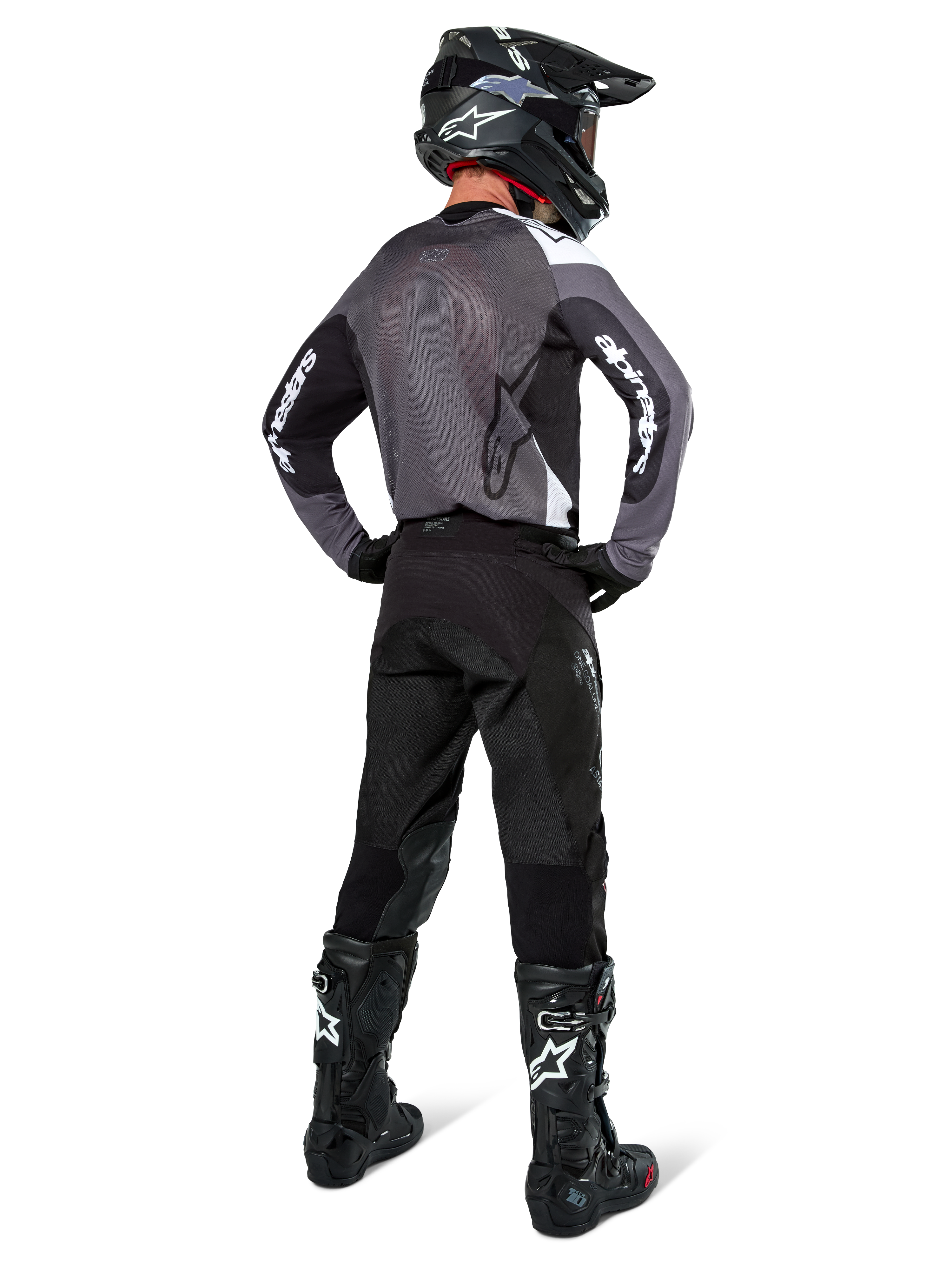 Motocross rider wearing Alpinestars Pro-Dura Jersey in Black Magnet White, rear view showing grey mesh back panel and black sleeves with white Alpinestars branding, coordinated with black pants, black helmet, and black and white boots, professional enduro racing gear