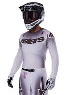 Alpinestars 2025 Supertech Maker Gear Set, Motocross-Komplettausrüstung in Weiß, Trikot mit grauem Farbverlauf, schwarzem Astars-Logo und Laser-Cut-Perforationen, passende Hose mit Magnetverschluss und Stern-Grafiken, schwarz-weißer Helm und Handschuhe für den Offroad-Racing-Look.